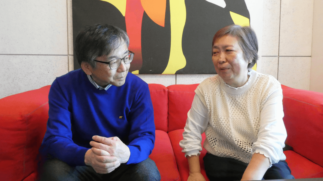 赤いソファに並んで座り、顔を見合わせながら話す飯塚正義さんと飯塚道代さんの画像