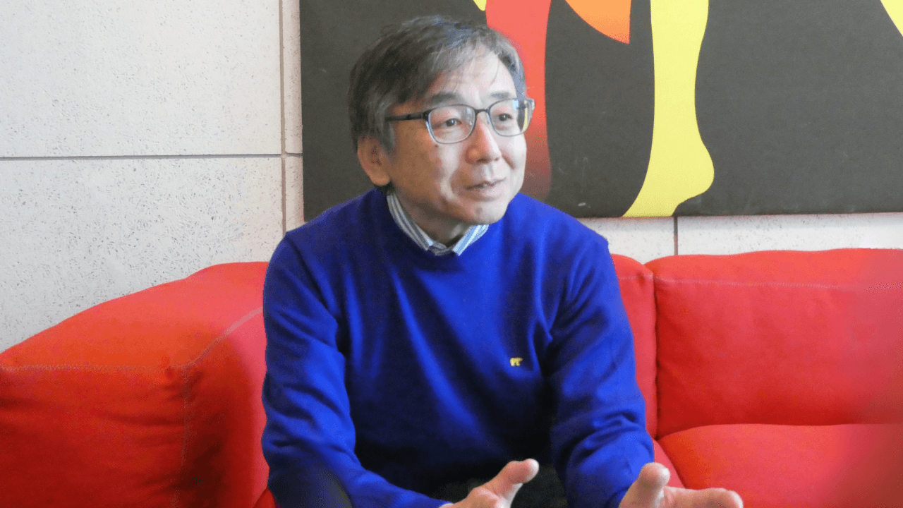 赤いソファに座り、手ぶりを交えながら話す飯塚正義さんの画像