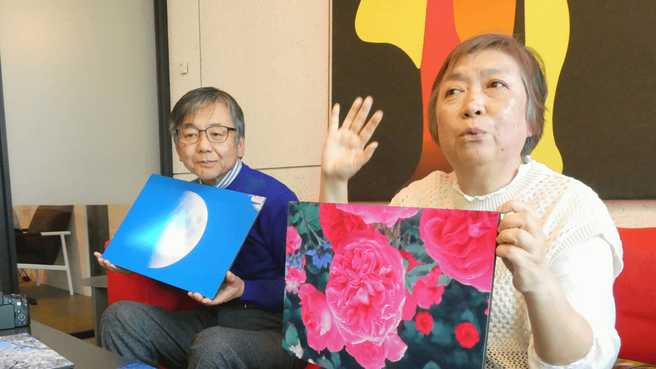 赤いバラの写真を手にする飯塚道代さんと、月の写真を手にする飯塚正義さんの画像