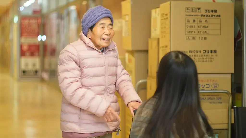 段ボールの並ぶ通路に杖を持って立つ大屋さん